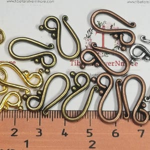 May include: A collection of 14 gold, silver, and copper-toned metal clasps. The clasps are shaped like a teardrop with a loop at the top. The clasps are arranged in a random pattern on a white surface. The clasps are approximately 1 inch in length.