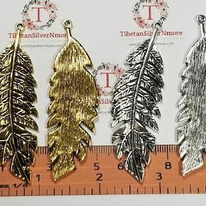 May include: Three metal feather charms in gold, silver, and antique silver. The charms are approximately 2 inches long and have a textured surface. They are shown on a ruler for size reference.