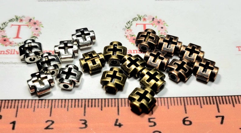 Puede incluir: Primer plano de dos pilas de cuentas de metal en forma de cruz. Una pila es plateada y la otra es una mezcla de bronce y cobre.
