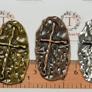 May include: Three oval-shaped metal charms with a cross design. The charms are in different colors: gold, copper, and silver. Each charm has a textured surface and a loop at the top for hanging.