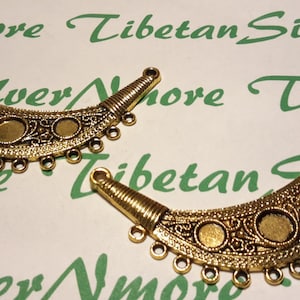 Puede incluir: Dos componentes de joyería en forma de media luna de color oro antiguo. Cada pieza presenta un diseño de filigrana detallado, tres engastes circulares y pequeños bucles a lo largo del borde inferior. El texto "Nmore Tibetan Silver" es visible en el fondo.