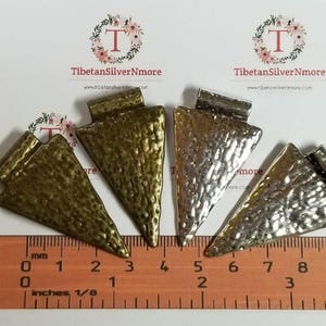 May include: Four silver and brass triangle-shaped pendant charms with a hammered texture. The charms are approximately 4 cm long and are arranged on a ruler with inch and centimeter markings.
