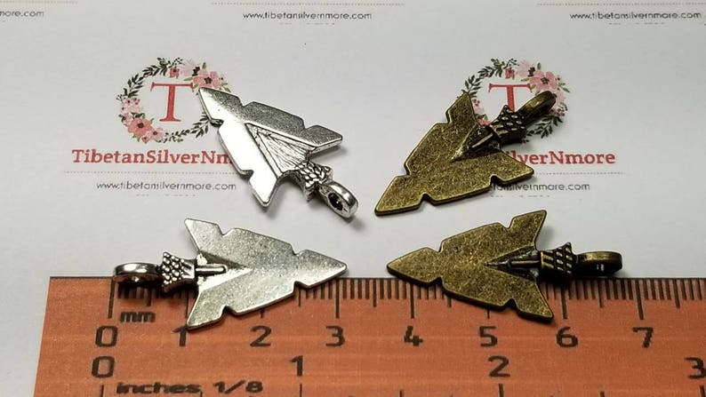 May include: Four arrowhead charms, two silver and two bronze, with a ruler for size reference. The charms are approximately 2 inches long.