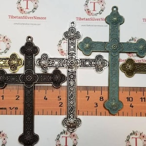 May include: Four ornate metal cross charms in gold, silver, black, and blue-green. The charms are approximately 2 inches in length and feature intricate designs. They are displayed on a ruler for size reference.