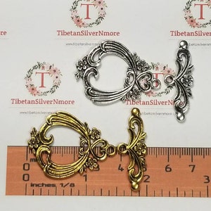 May include: Two ornate metal connector charms, one gold and one silver, with a floral design. The charms are approximately 1 inch in length and are shown next to a ruler.
