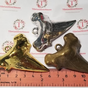 Könnte beinhalten: Drei Hai-Zahn-Anhänger in verschiedenen Ausführungen: Gold, Silber und Bronze. Jeder Anhänger hat eine Öse zum Anbringen an einer Halskette oder einem Armband. Die Anhänger werden auf einem Lineal in Zoll gemessen.