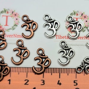 May include: A collection of silver and copper Om symbol charms. The charms are about 1 inch in size and have a hole at the top for hanging. The charms are arranged on a white surface with a ruler for scale.