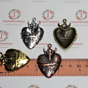 Puede incluir: Cuatro colgantes en forma de corazón de color plateado, dorado y cobre con una cruz y espinas. Los colgantes miden aproximadamente 5 cm y se muestran en una regla.