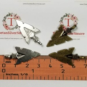 May include: Four silver and bronze arrowhead charms with a loop for attaching to jewelry or other crafts. The charms are shown on a ruler with measurements in inches.