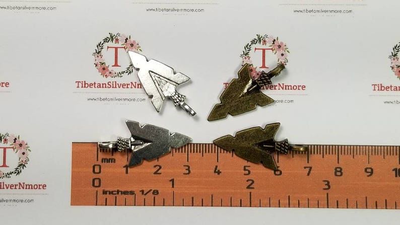 May include: Four silver and bronze arrowhead charms with a loop for attaching to jewelry or other crafts. The charms are shown on a ruler with measurements in inches.