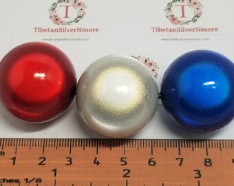 Könnte beinhalten: Drei große, runde Perlen auf einer schwarzen Schnur. Die Perlen sind rot, silber mit goldenem Schimmer und blau. Die Perlen haben einen Durchmesser von etwa 5 cm.