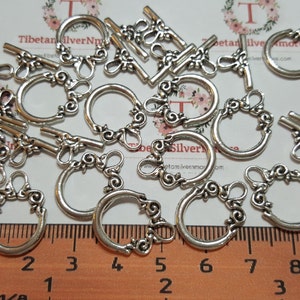 May include: A collection of silver-colored toggle clasps and round clasps with decorative scrollwork. The clasps are arranged on a surface with a ruler showing inches and millimeters. The clasps are likely for jewelry making or crafts.