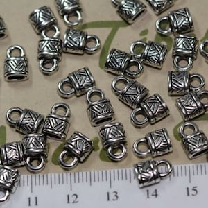 May include: A close-up of a group of silver metal end caps with a decorative pattern. The end caps have a loop on one end and a closed end on the other. The end caps are arranged on a white surface with a ruler in the background.