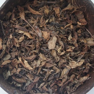 May include: A close-up shot of dried leaves in a brown coconut bowl. The leaves are various shades of brown and tan, with some darker areas. The bowl has a natural, textured appearance, suggesting a rustic or organic theme.