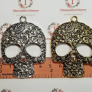 May include: Two silver and gold metal skull charms with intricate floral designs. The charms are approximately 2 inches in length and have a loop at the top for hanging.