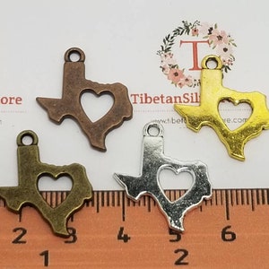 May include: Four metal charms in the shape of the state of Texas, each with a heart cutout. The charms are in bronze, gold, silver, and antique brass colors. The image includes the text "TibetanSilverNmore".
