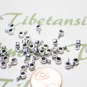May include: A collection of small, silver-colored jewelry findings, including crimp ends and bails, scattered on a light-colored surface. The findings are round with a loop for attaching to other jewelry components. A penny is included for size reference.