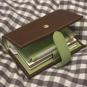 May include: A brown and green leather planner with a snap closure. The planner is open, revealing a green interior with a zippered pocket and several notepads. The stitching is white, and the hardware is gold.