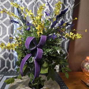 May include: A floral arrangement featuring yellow and purple flowers, green foliage, and a purple velvet bow. The arrangement is set on a wooden base with a decorative floral design. The background includes a patterned curtain.