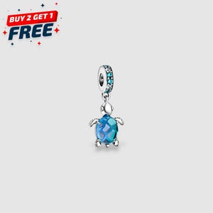 Puede incluir: Un charm de plata con un diseño de tortuga marina azul y turquesa. El charm tiene una parte superior redonda con piedras azules y turquesas. La tortuga tiene un caparazón azul y turquesa. La imagen también incluye el texto "BUY 2 GET 1 FREE".