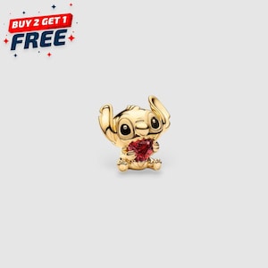 May include: A gold-colored Stitch charm holding a red heart-shaped gem. The charm has large, expressive eyes and is set against a light background. Text in the upper left corner reads "BUY 2 GET 1 FREE".