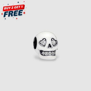 May include: A white skull charm with heart-shaped eye sockets and a row of sparkling stones for teeth. The charm has a black hole on the side and is likely made of metal. The image also includes the text "BUY 2 GET 1 FREE".