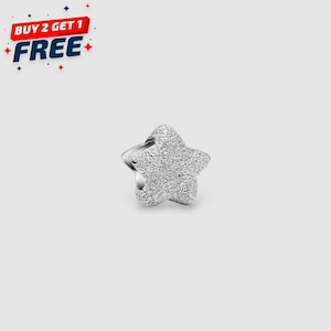 Puede incluir: Un charm plateado en forma de estrella con una superficie texturizada. El charm tiene un anillo en la parte posterior. La imagen también incluye el texto "BUY 2 GET 1 FREE" en rojo, blanco y azul.