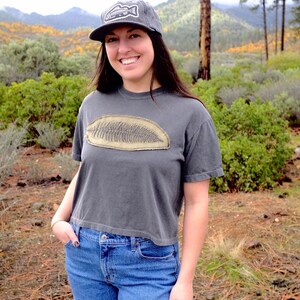 May include: A gray t-shirt with a large, tan fossil design. The person is wearing a gray baseball cap with a fish logo and blue jeans. The background features a scenic view of mountains and trees.