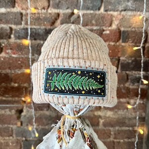 May include: Beige knit beanie with a rectangular patch featuring a green fern design on a black background, framed by a light blue border with yellow stars and crosses. The beanie is displayed against a brick wall.