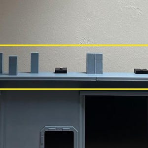 May include: A light blue and grey model building featuring a series of small, rectangular structures on its roof. These structures vary in shape and size, with small, black rectangular objects also present.