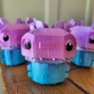 Peut inclure: Un groupe de petites figurines imprimées en 3D représentant une créature au design cubique et stylisé. Chaque figurine possède une partie supérieure rose vif avec de grands yeux noirs et une base bleue. La texture montre des couches horizontales.