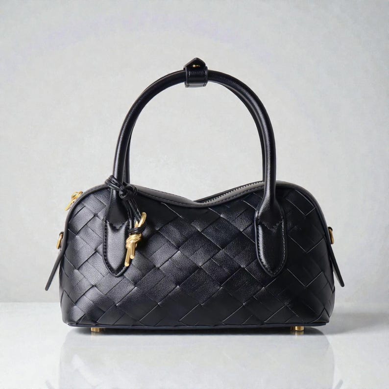 May include: Black leather handbag with a woven pattern. The bag has a rounded shape, a top handle, and a gold-colored key charm. The zipper and base studs are also gold. The bag is sitting on a white surface.