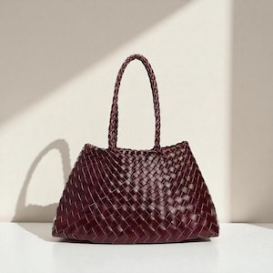 Peut inclure: Un sac fourre-tout en cuir bordeaux tressé avec une poignée tressée. Le sac a une forme trapézoïdale et un design structuré. Le motif de tissage est serré et uniforme, créant une surface lisse. Le sac est posé sur un fond neutre.