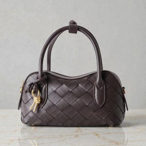 May include: A dark brown handbag with a woven pattern. The bag has two handles and a small top handle. A gold-colored charm hangs from the side. The bag is a classic style, suitable for everyday use.