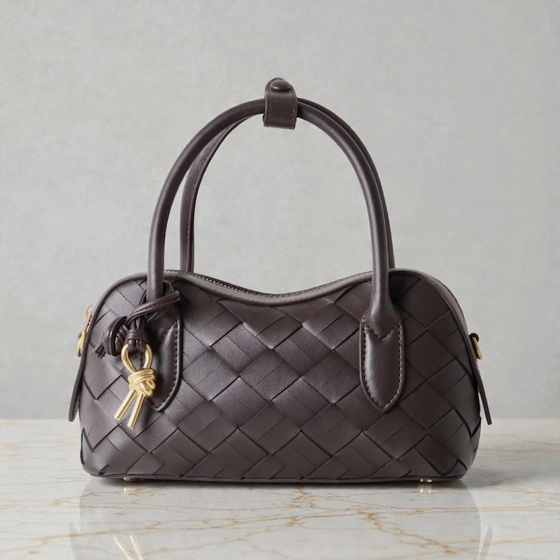 May include: A dark brown handbag with a woven pattern. The bag has two handles and a small top handle. A gold-colored charm hangs from the side. The bag is a classic style, suitable for everyday use.