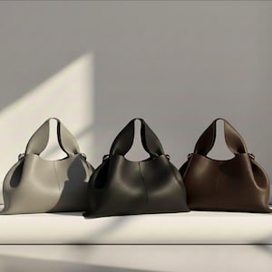 May include: Three leather tote bags in a studio setting. The bags are in shades of gray, black, and brown. Each bag has a rounded shape with a top handle. The bags are sitting on a white surface against a white background.