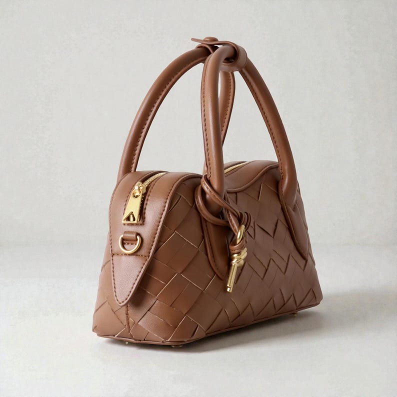 May include: A brown leather handbag with a woven pattern. The bag has two short handles and a gold zipper closure. A gold-colored key-shaped charm hangs from the handle. The bag is a trapezoid shape.