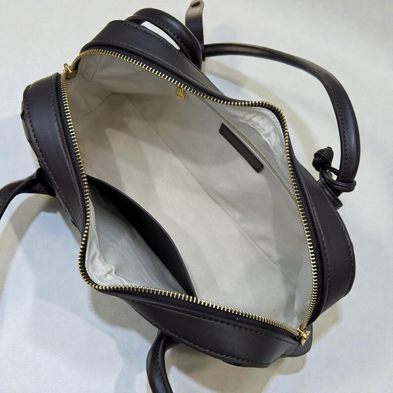May include: Dark brown leather handbag with a gold zipper and handles. The bag is open, revealing a white interior with a pocket. The handles are knotted at the top. The bag is a satchel style.