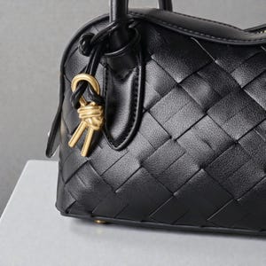 May include: Black leather handbag with a woven pattern. The bag features a top handle and a gold-colored charm. The leather has a textured appearance, and the bag appears to be new.