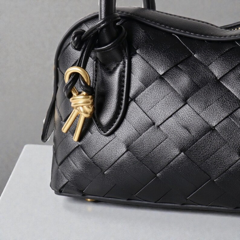 May include: Black leather handbag with a woven pattern. The bag features a top handle and a gold-colored charm. The leather has a textured appearance, and the bag appears to be new.
