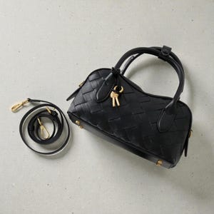 May include: Black woven leather handbag with a top handle and a detachable shoulder strap. The bag features gold-tone hardware, including a key-shaped charm and small feet on the base. The bag's design is a structured, barrel shape.