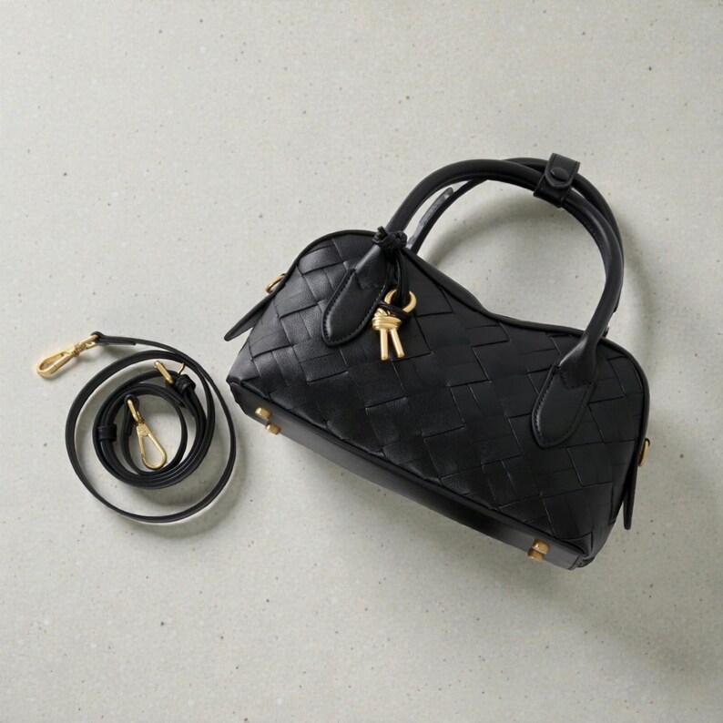 May include: Black woven leather handbag with a top handle and a detachable shoulder strap. The bag features gold-tone hardware, including a key-shaped charm and small feet on the base. The bag's design is a structured, barrel shape.