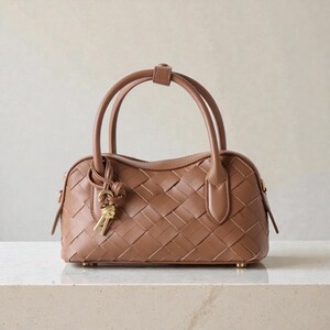 May include: A brown woven handbag with two top handles and a small handle in the middle. The bag has a rectangular shape with a gold-colored key charm hanging from the side. The bag is sitting on a white surface.