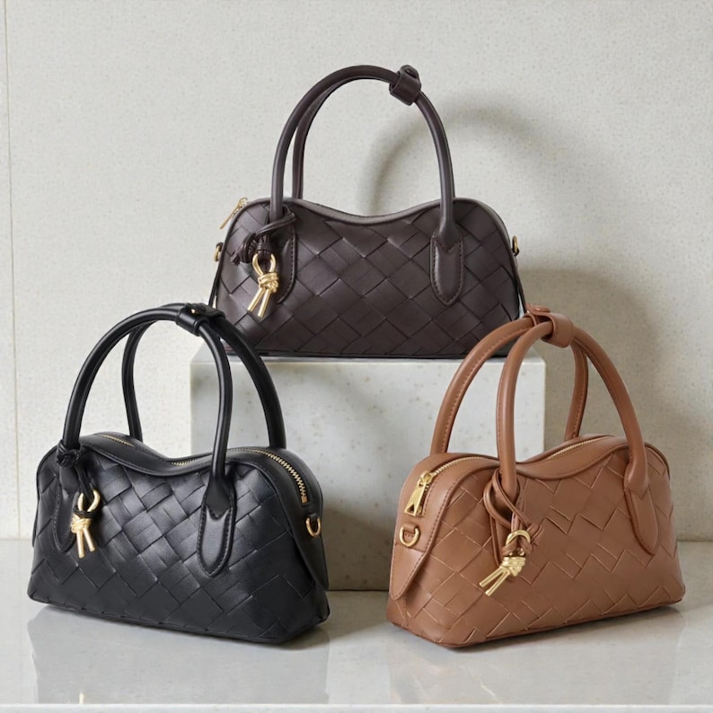 May include: Three woven leather handbags in black, brown, and tan. Each bag features a top handle, a gold zipper, and a decorative gold key charm. The bags are displayed on a white surface.
