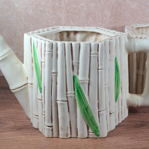 Op de afbeelding: Off-white keramische gieter en bijpassende plantenbak. De gieter heeft een tuit en bamboe-achtige details. De plantenbak heeft een handvat en bamboe-achtige details. Groene bladaccenten zijn op beide stukken geschilderd. De stukken staan op een houten oppervlak.