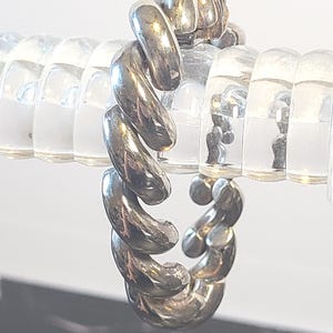 May include: A silver-colored ring with a twisted, rope-like design. The ring's metal has a polished, reflective surface. The ring is displayed against a clear, coiled background.