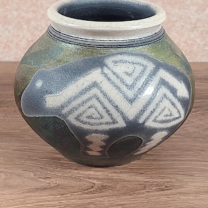 May include: A handcrafted ceramic vase with a rounded body and a wide opening. The vase features a bear design in white and gray, set against a backdrop of green and blue hues. The rim is a light beige color.