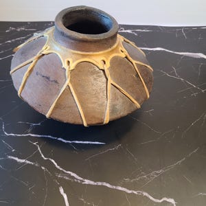 Könnte beinhalten: Eine handgefertigte, braune Keramikvase mit dunklem Innenraum und einem hellbraunen, netzartigen Design an der Außenseite. Die Vase hat einen abgerundeten Körper und eine weite, offene Mündung. Das dekorative Stück steht auf einer schwarzen Marmoroberfläche.