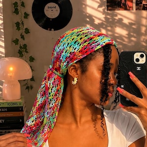 Può includere: Un foulard all'uncinetto multicolore caratterizzato da un vivace motivo a maglia aperta nei toni del rosa, blu, giallo e verde. Il copricapo triangolare è annodato sul retro sopra i capelli scuri e abbinato a una t-shirt bianca a maniche corte.