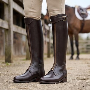 Könnte beinhalten: Dunkelbraune Lederreitstiefel mit durchgehenden Reißverschlüssen und goldfarbenen Schnallen. Die Stiefel werden mit beigefarbenen Reithosen getragen. Im Hintergrund ist ein Pferd mit Sattel zu sehen.